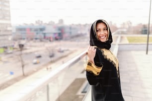 Schöne muslimische Frau mit zahnigem Lächeln und Schal auf dem Kopf, die auf dem Dach posiert.