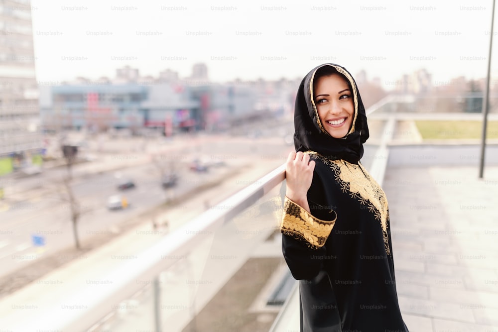 이빨 미소와 머리에 스카프를 두른 아름다운 무슬림 여성이 옥상에서 포즈를 취하고 있다.