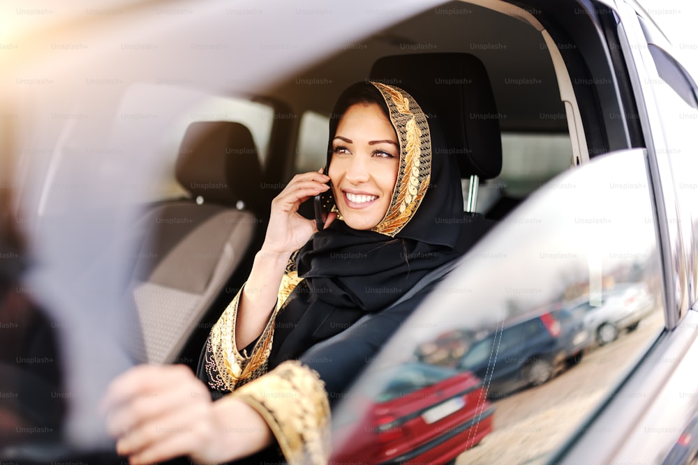 Belle femme musulmane avec un sourire aux dents et un foulard sur la tête parlant au téléphone et tenant la main sur le volant.