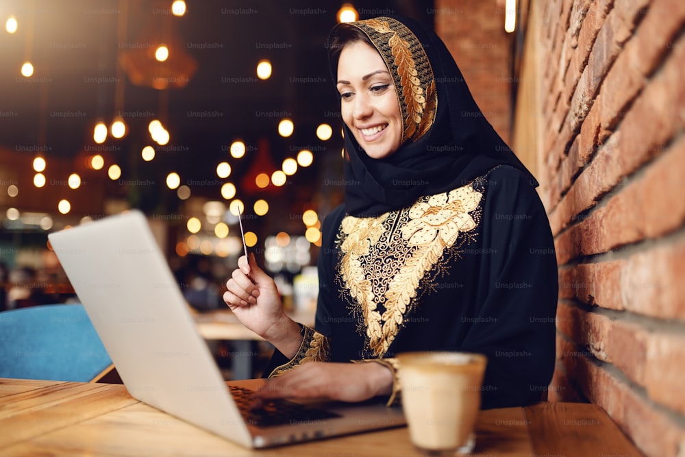 Gorgeous muslim woman with toothy smile dressed in traditional wear using laptop for on-line shopping while sitting in cafeteria. On desk coffee.