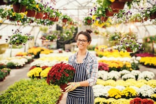 Happy Caucasian female florist holding flowers in pot while standing in greenhouse.