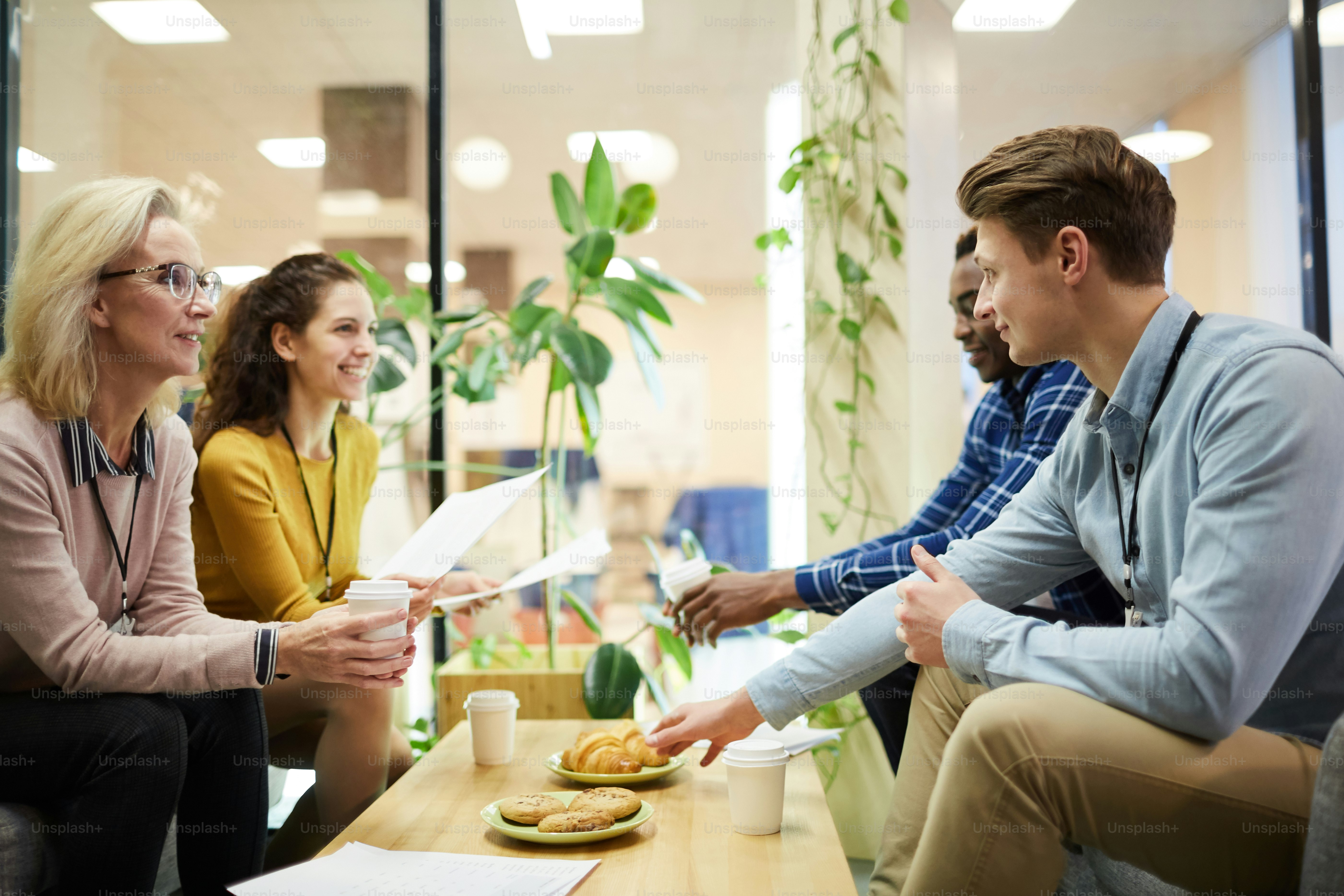 Four intercultural colleagues sitting by table and discussing papers while having coffee with pastry in office