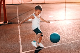 셔츠 아래 모래 반바지를 입은 어린 소년이 아침에 코트에서 공을 차고 있다.