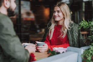 Waist up portrait of charming girl sitting at the table and holding cup of coffee. She looking at bearded man and smiling