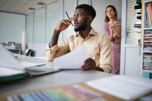 Jeune homme d’affaires pensif avec du papier et un crayon assis près du bureau tandis que son collègue franchit la porte du bureau derrière son dos