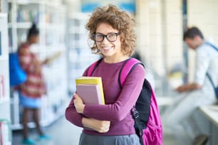 Hermosa estudiante segura de sí misma con gafas sosteniendo libros y mirando a la cámara mientras está parada en el pasillo en las vacaciones de la escuela secundaria