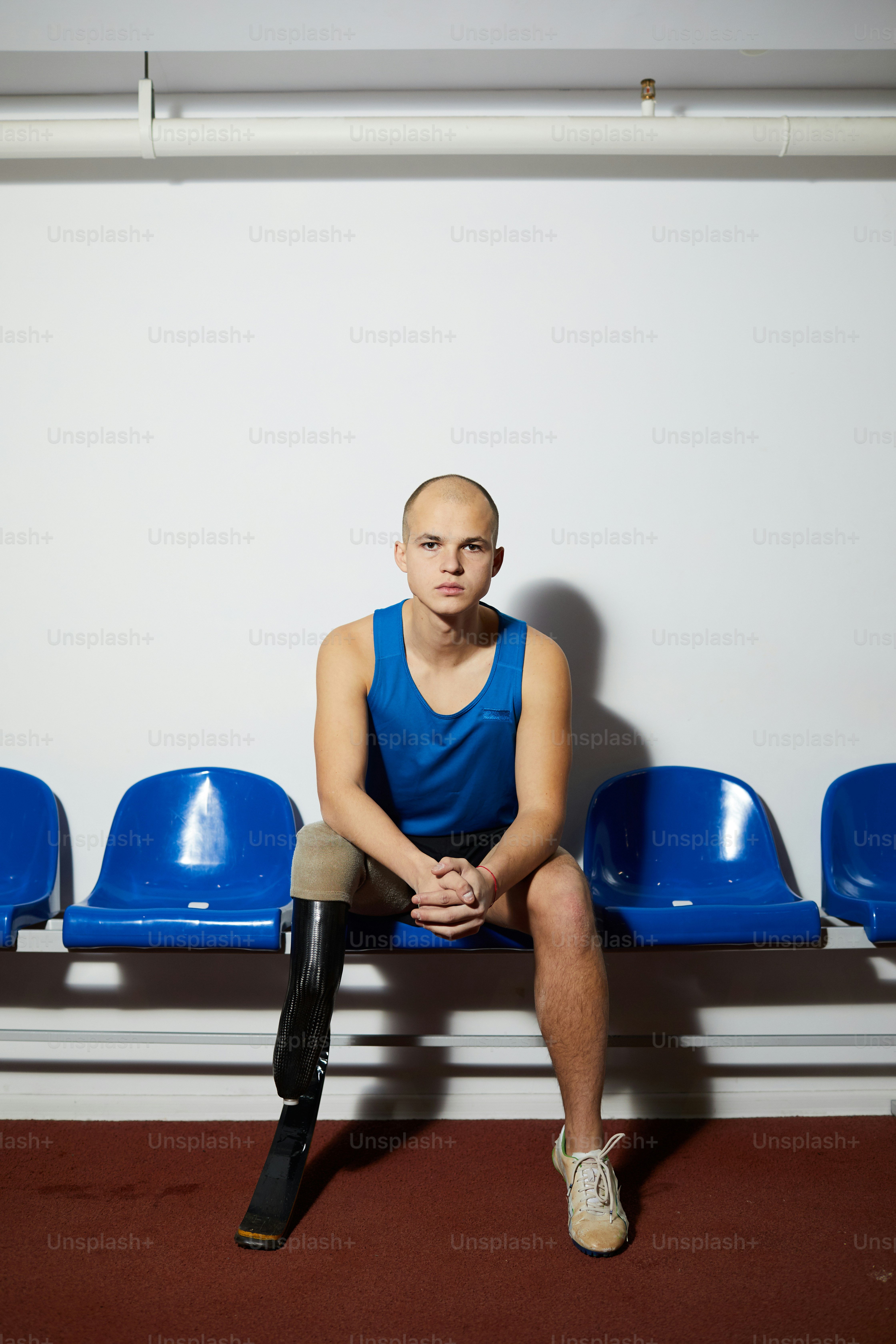 Behinderter junger Sportler mit Handicap, der vor dem Wettkampf auf einem blauen Plastikstuhl an der Wand sitzt