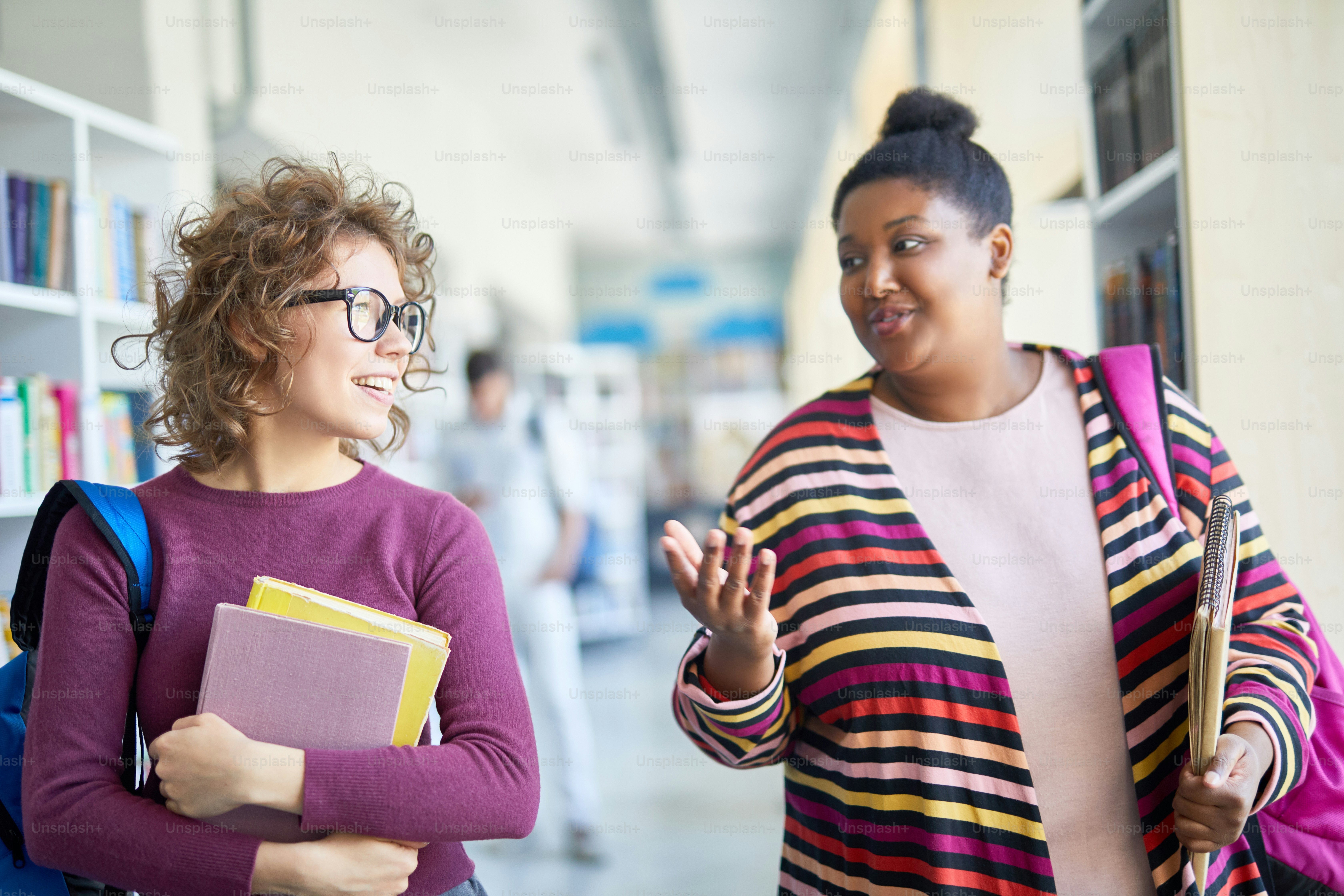 Positive friendly interracial students in bright clothing chatting while walking over corridor: confident African-American girl gesturing while explaining her idea to groupmate
