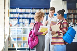 Cheerful confident multi-ethnic student friends with colorful satchels standing in circle and discussing literature in library