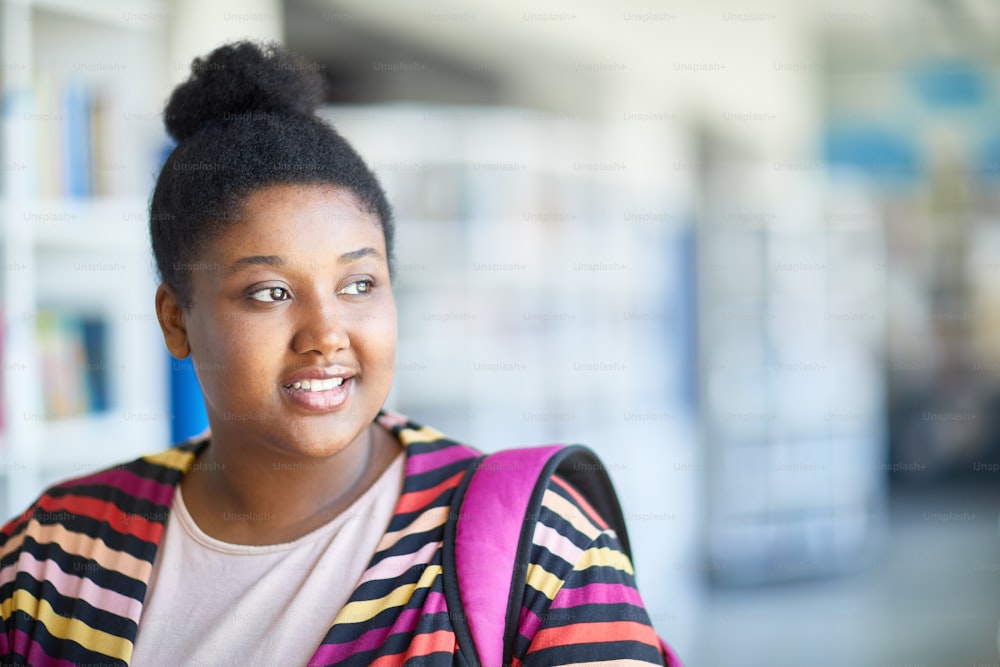 Sonriendo atractiva estudiante afroamericana con sobrepeso con moño de pelo reflexionando sobre el futuro y mirando a un lado mientras piensa en el ingreso a la universidad