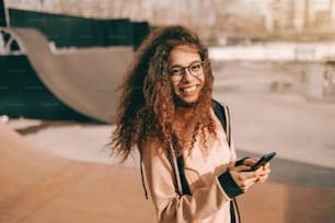 스케이트 공원에 서 있는 동안 스마트폰을 사용하여 이빨 미소를 짓는 예쁜 혼혈 십대 힙스터 소녀의 클로즈업.