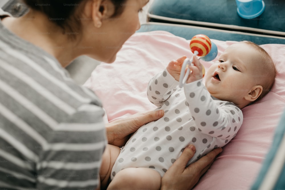 Foto ritagliata della madre che guarda il suo bambino appena nato. Il bambino sta giocando con il sonaglio mentre è sdraiato sul divano di casa. Concetto di maternità felice