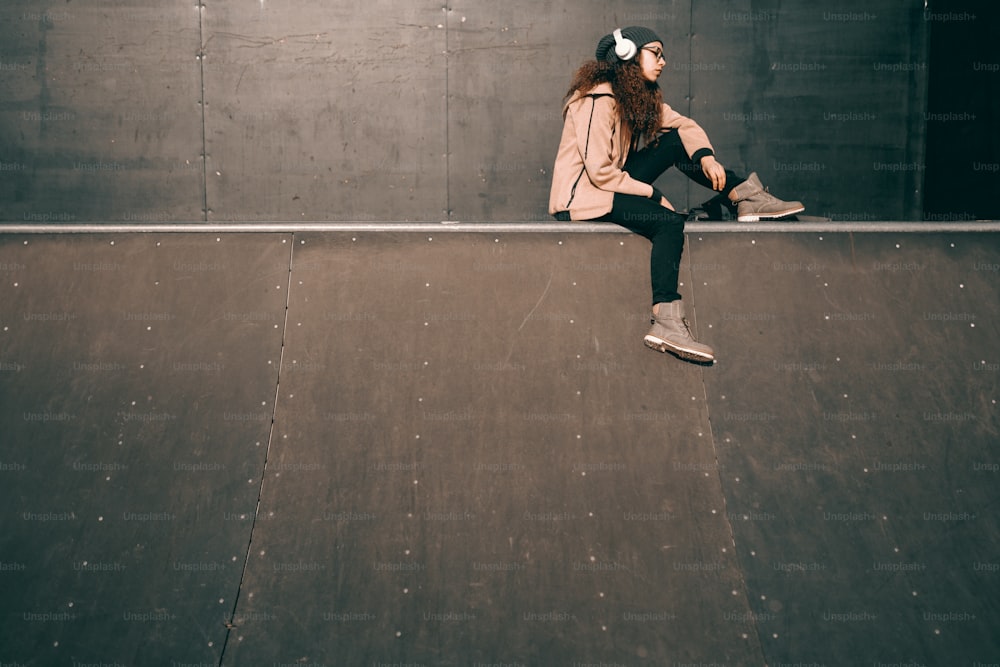 Adolescente hipster métisse avec une expression faciale sérieuse appréciant la musique et assise dans un skate park.