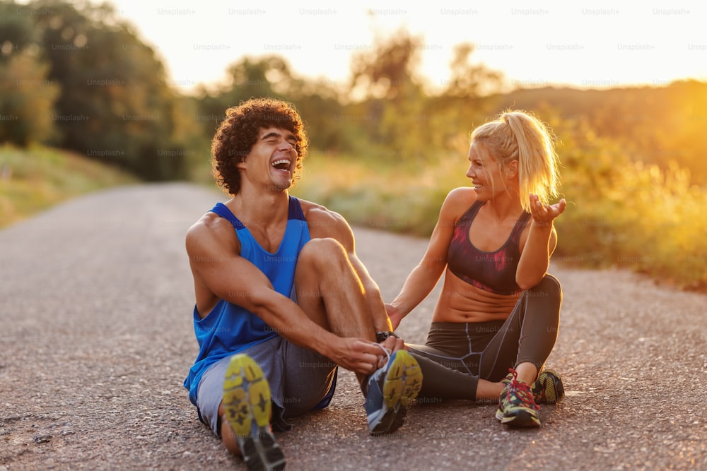 Fit cute couple sitting on the country road and resting from running. Man tying shoelace. Summer time in the morning.