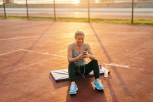 Smiling blonde Caucasian woman sitting on the court with earphones in ears and smart phone in hands in the morning.