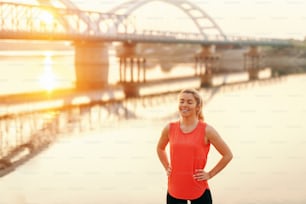 Smiling Caucasian sporty woman with hands on hips resting from running. In background river, bridge and morning sun.
