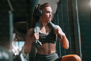 Joven atractiva culturista musculosa fuerte con cola de caballo y auriculares posando en gimnasio con toalla alrededor del cuello. Los sueños no funcionan a menos que tú lo hagas.