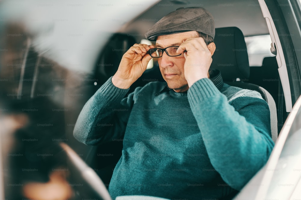 Caucasian senior man with cap on head putting eyeglasses while sitting in a car. Window opened.