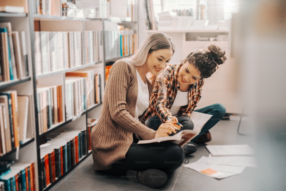 Two Caucasian pretty female students sitting with legs crossed in library and studying for exams. Girl with curly hair pointing at answer in book.