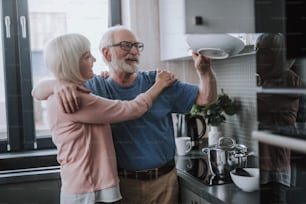 Enjoying to make household duties together. Waist up portrait of happy senior woman and man embracing while taking plate from wall hanging cupboard on kitchen