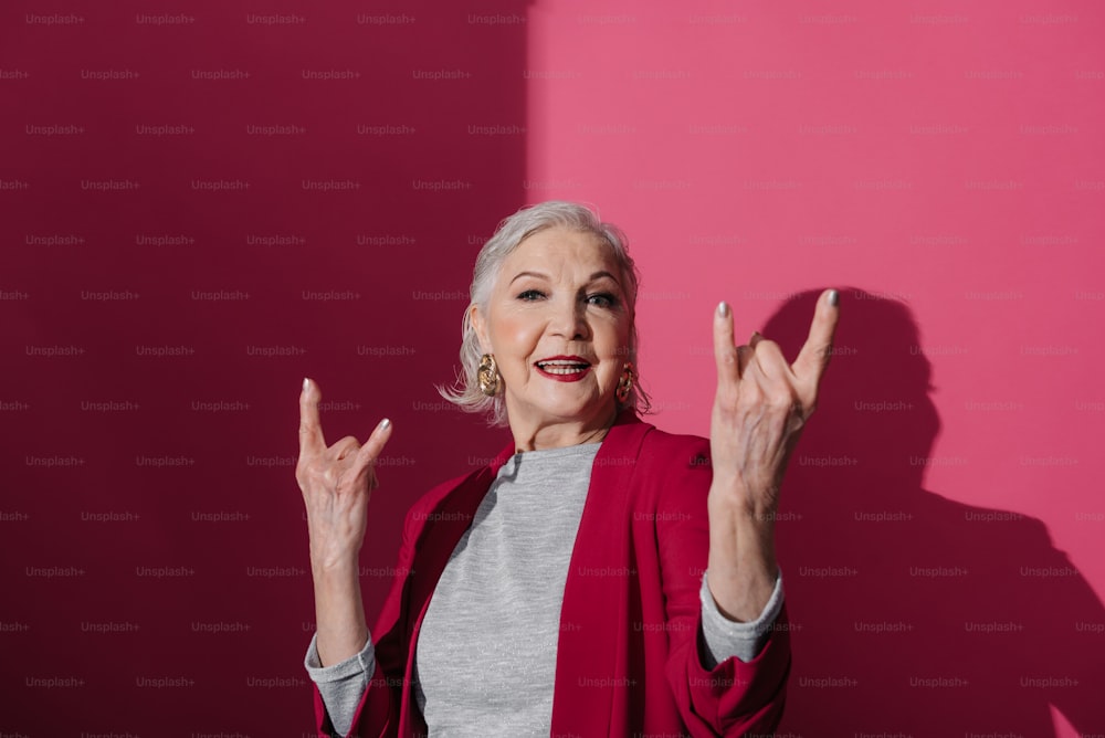 Waist up of senior stylish woman wearing modern bright jacket isolated on red background and showing rock horns. She is posing and looking at camera