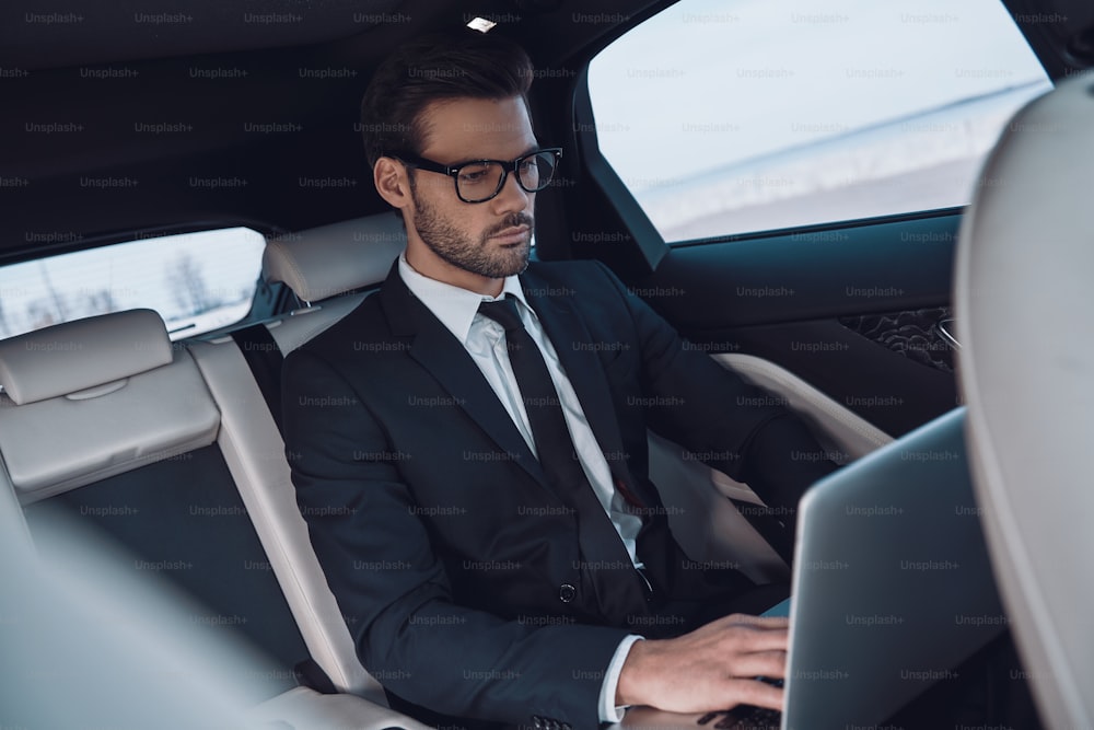 Hübscher junger Mann in vollem Anzug, der mit dem Laptop arbeitet, während er im Auto sitzt