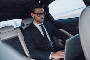 Beau jeune homme en costume complet travaillant à l’aide d’un ordinateur portable assis dans la voiture
