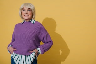 Cropped photo of confident stylish old lady putting her hands in pockets while situating against yellow wall. She is wearing fashionable clothes. Copy space in right side