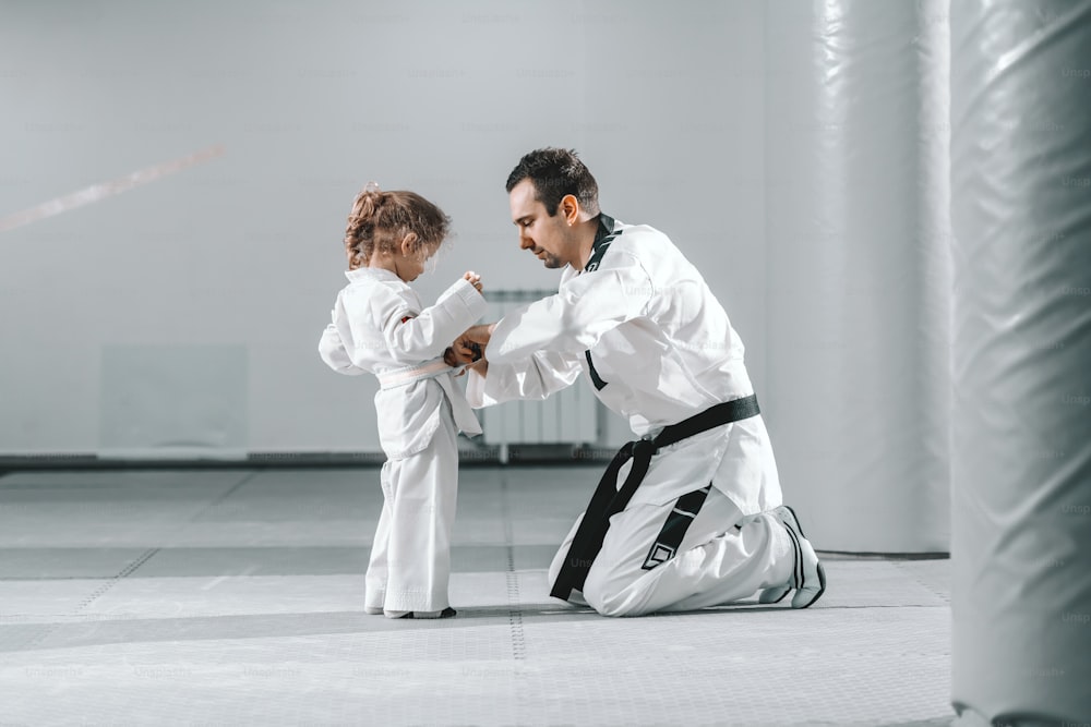 Taekwondo trainer kneeling and tying belt to little cute girl with curly hair.