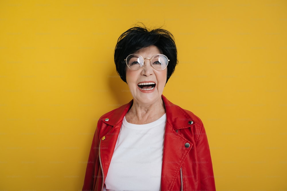 Cropped photo of of stylish happy woman wearing modern red jacket and cute eyeglasses while looking at camera on yellow background. She is laughing with opened mouth while posing indoors