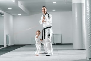 Lächelnder kaukasischer Taekwondo-Trainer posiert mit kleinem Mädchen in weißer Turnhalle.