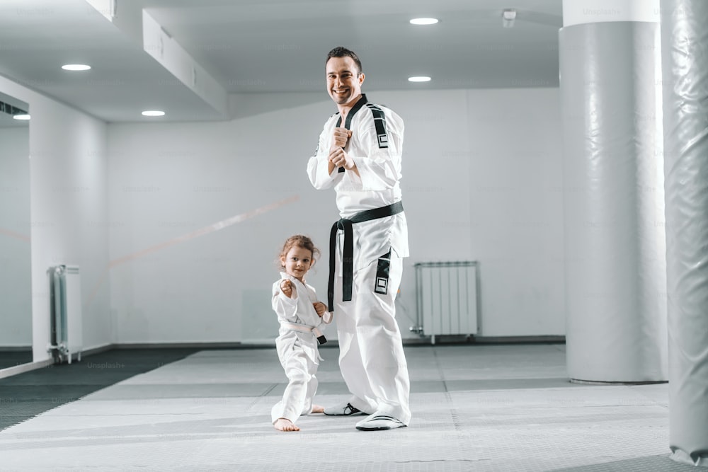 Smiling Caucasian taekwondo trainer posing with little girl in white gym.
