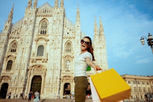 happy stylish solo traveller woman in white t-shirt and sunglasses rejoicing walking with shopping bags in the front of Duomo in Milan, Italy.