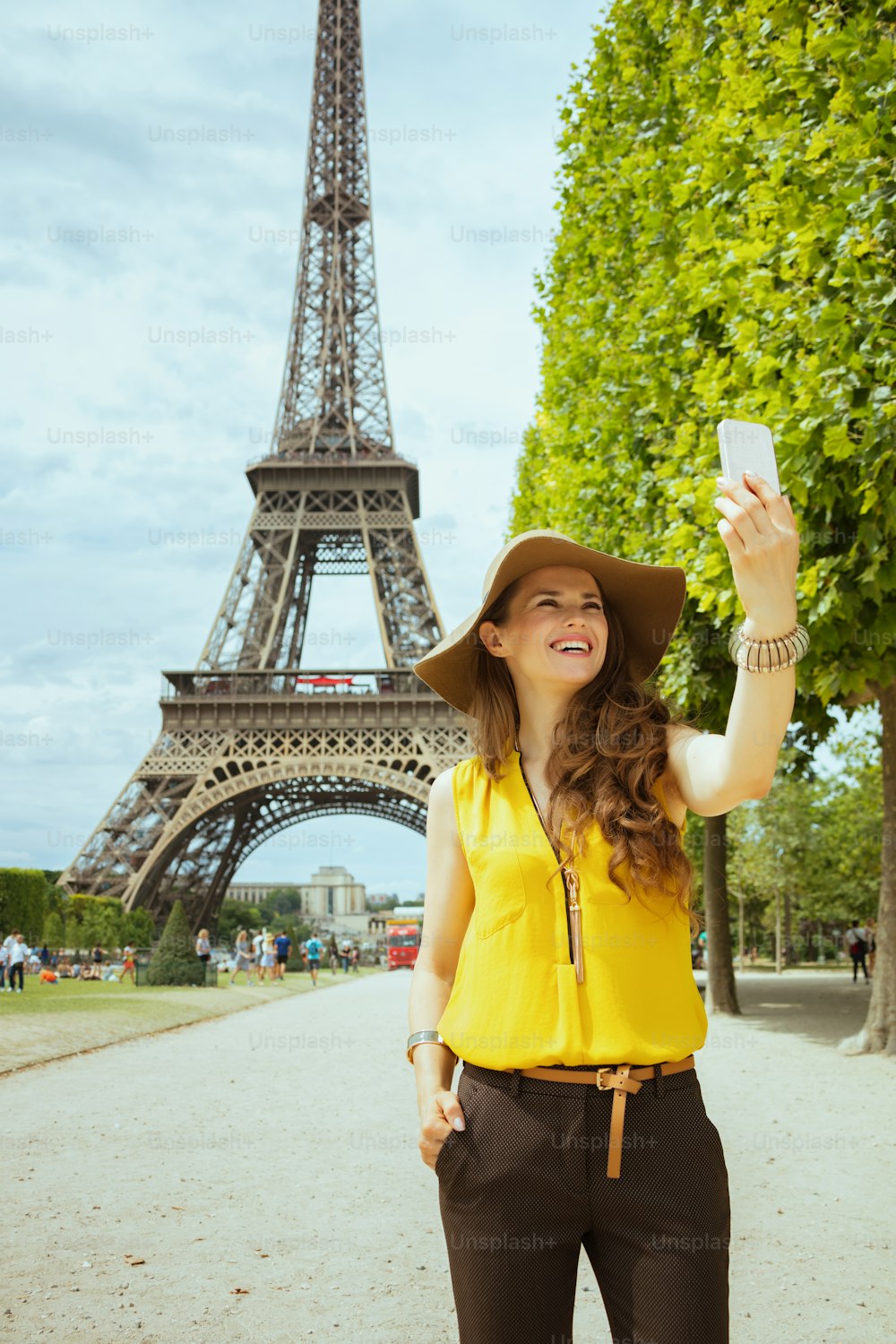 フランスのパリのエッフェル塔に対してスマートフォンで自分撮りをする黄色いブラウスと帽子をかぶった幸せな現代の一人旅の女性。