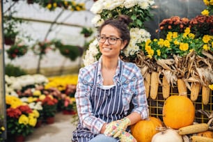 Charming Caucasian florist in apron and gardening gloves on sitting in front of greenhouse