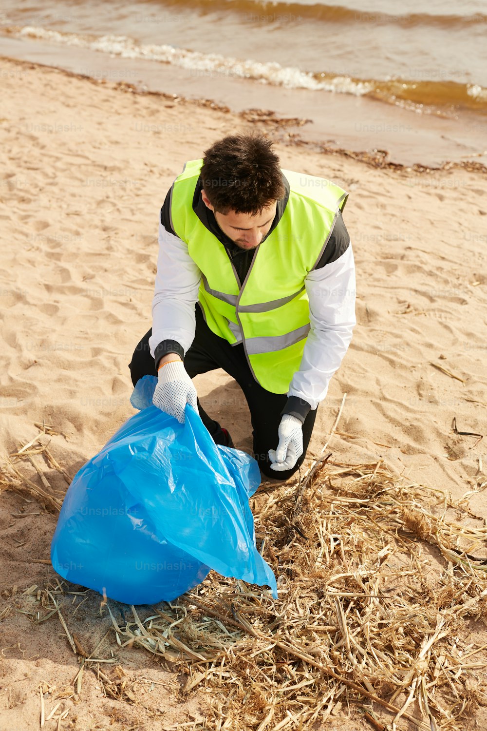 Joven voluntario contemporáneo en uniforme recogiendo desechos mientras trabaja en una playa de arena
