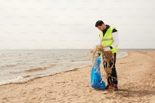 Junge Freiwillige in schützender Arbeitskleidung, die am Sandstrand stehen, während sie während der Arbeit natürliche Abfälle in einen blauen Sack werfen