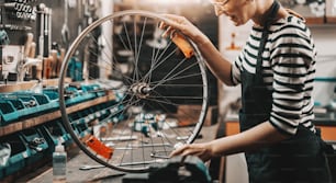 Cute Caucasian female worker holding and repairing bicycle wheel while standing in bicycle workshop.