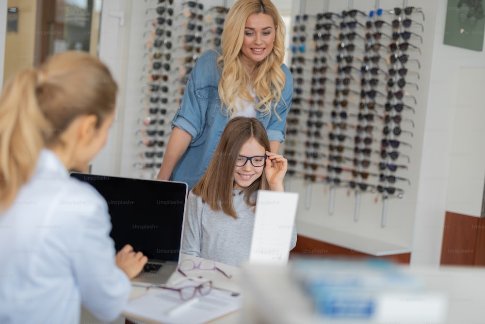 Angenehme, fürsorgliche Mutter, die mit ihrer Tochter ein Optikgeschäft besucht und gleichzeitig eine Brille für ein besseres Sehvermögen wählt