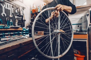 Smiling handsome Caucasian man holding bicycle wheel in hands while standing in workshop.