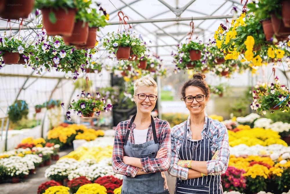 Two Caucasian florists in aprons standing in greenhouse with arms crossed surrounded by flowers.