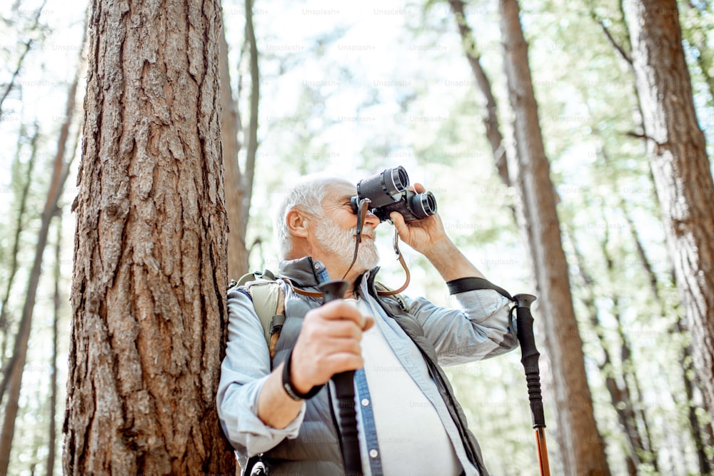 Portrait of a senior man with binoculars and backpack resting near the tree while traveling in the forest
