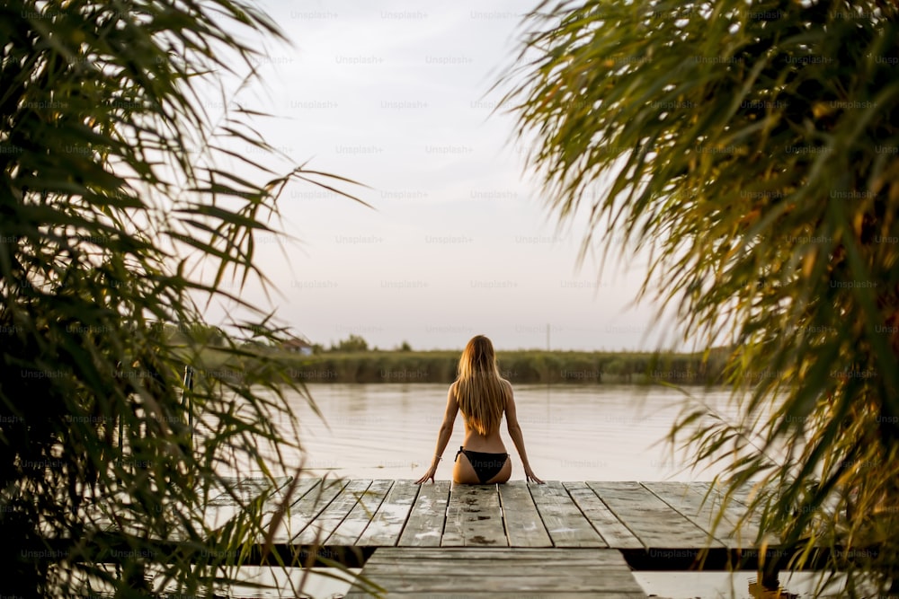 Vista posteriore alla giovane donna attraente in bikini che si siede su un molo al lago