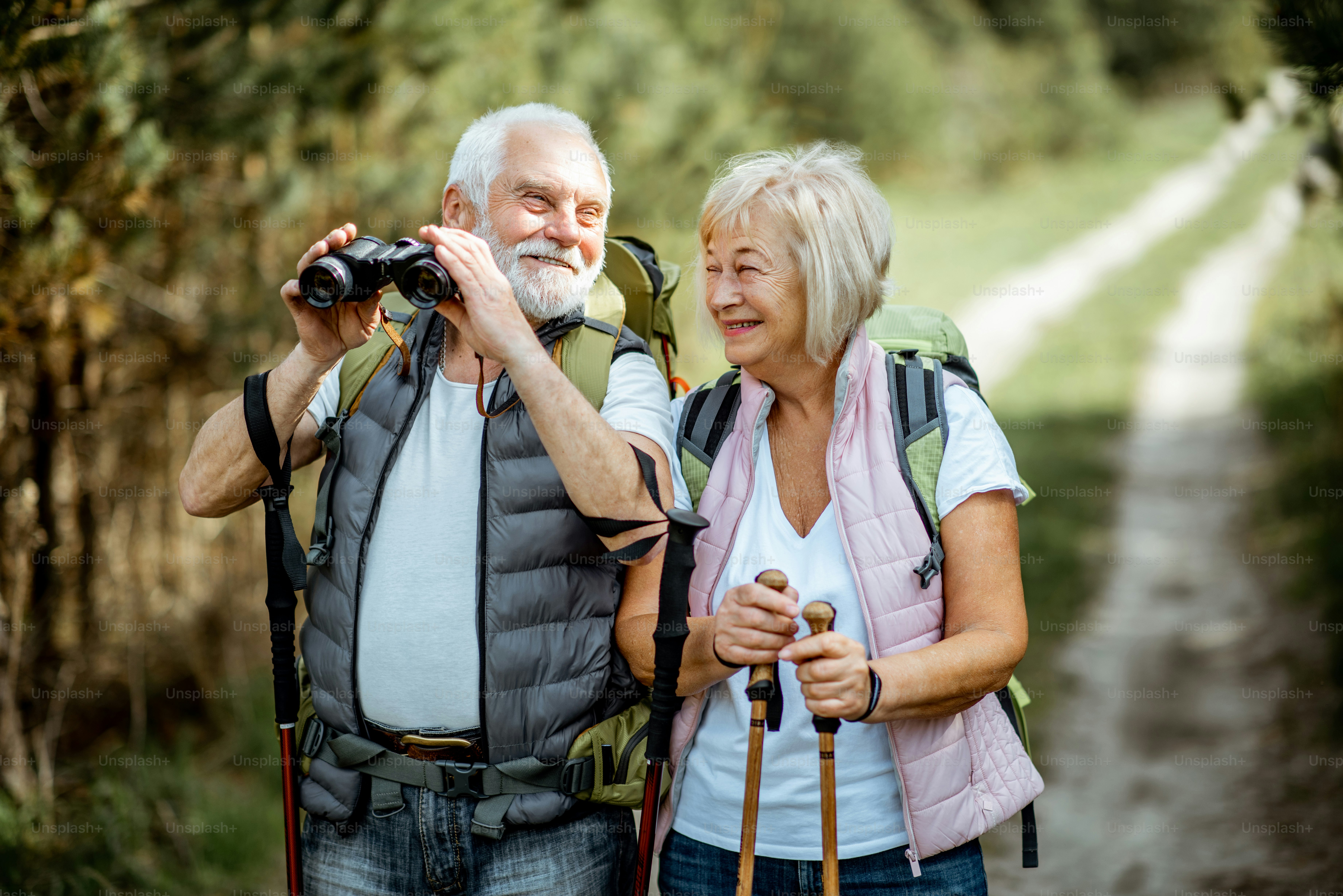 An older man with binoculars and a woman hiking on a forest path