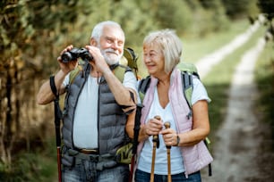 Portrait of a happy senior couple standing together with binoculars, backpacks and trekking sticks while hiking in the forest. Concept of an active lifestyle on retirement