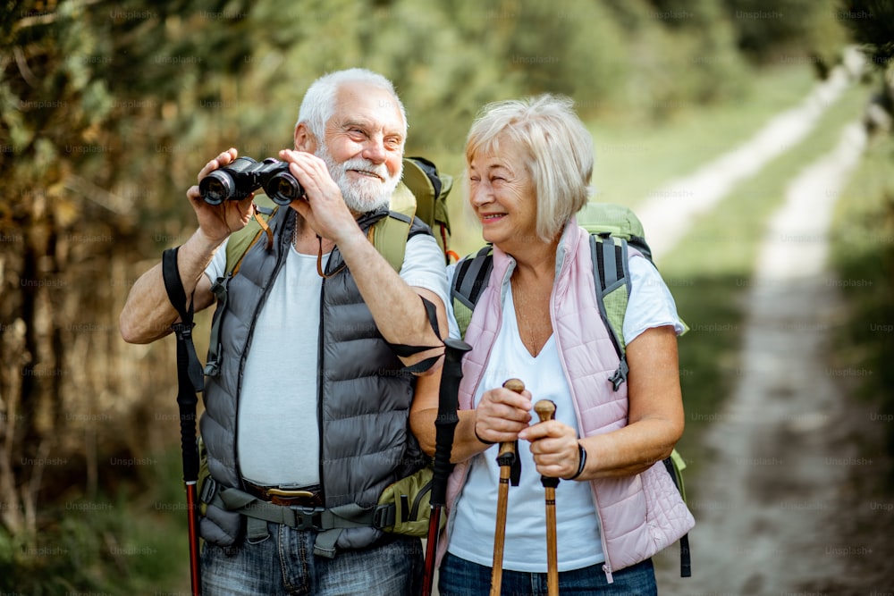 Porträt eines glücklichen Seniorenpaares, das mit Fernglas, Rucksäcken und Trekkingstöcken beim Wandern im Wald zusammensteht. Konzept eines aktiven Lebensstils im Ruhestand
