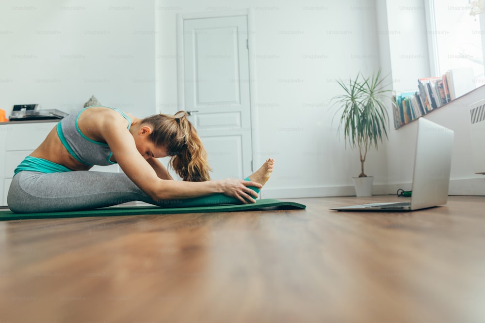 Health practice concept. Side on portrait of young lady in sportswear doing paschimottanasana yoga pose on mat in front of laptop. Seated forward bend exercise