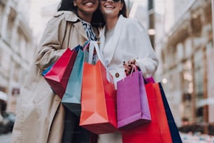Close up of smiling girls in trench coats holding colorful shopping bags