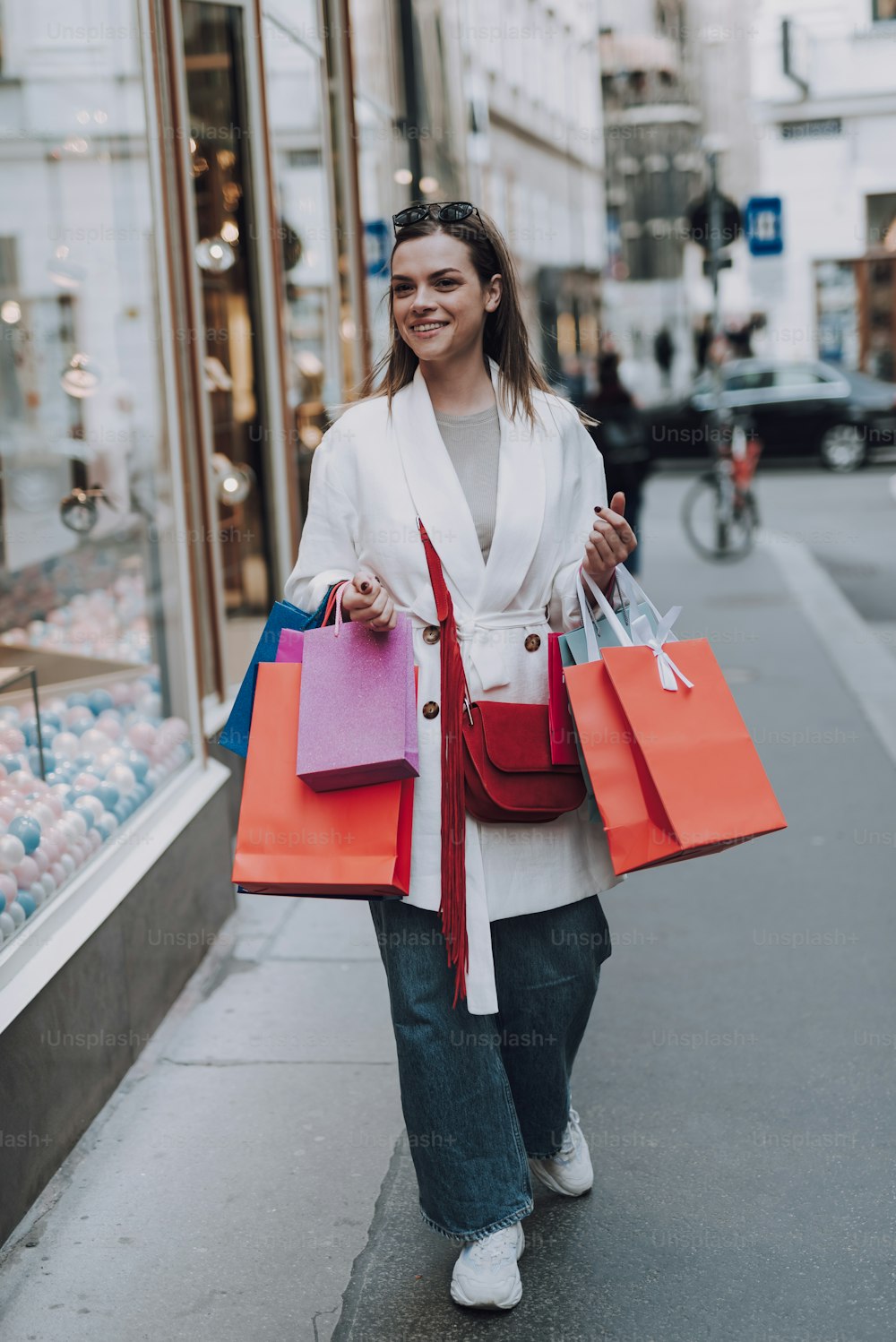 Full length portrait of charming stylish lady in trench coat holding colorful shopping bags. She looking away and smiling