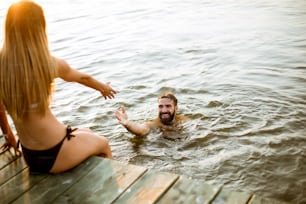 Giovani coppie felici che godono mentre la donna seduta sul molo e l'uomo che nuota nel lago